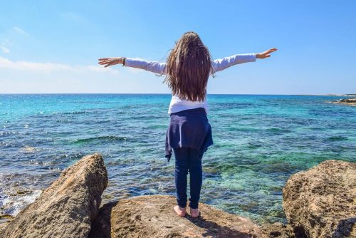 Girl with arms outstretched standing on rock by water